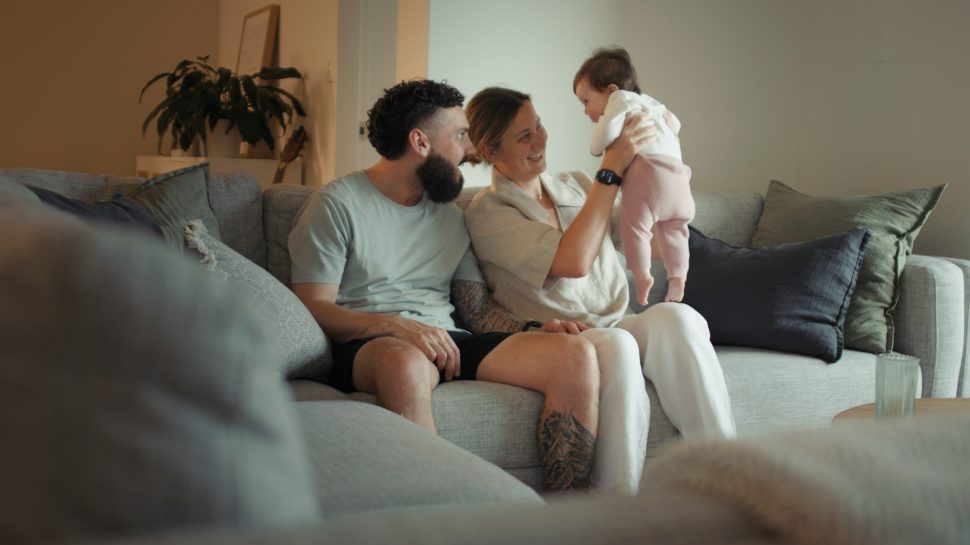 Andrew Eagers at home with his partner and baby daughter. Couple smiles as they hold their baby. 