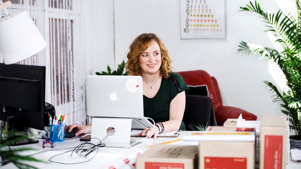 Helle is sitting at her work desk behind an Apple computer. To her left is a monitor, stationary and a lamp, to her right is more stationary, papers and a pot plant. IN the foreground of the image is Australia Post branded mailing boxes and cylinders.  