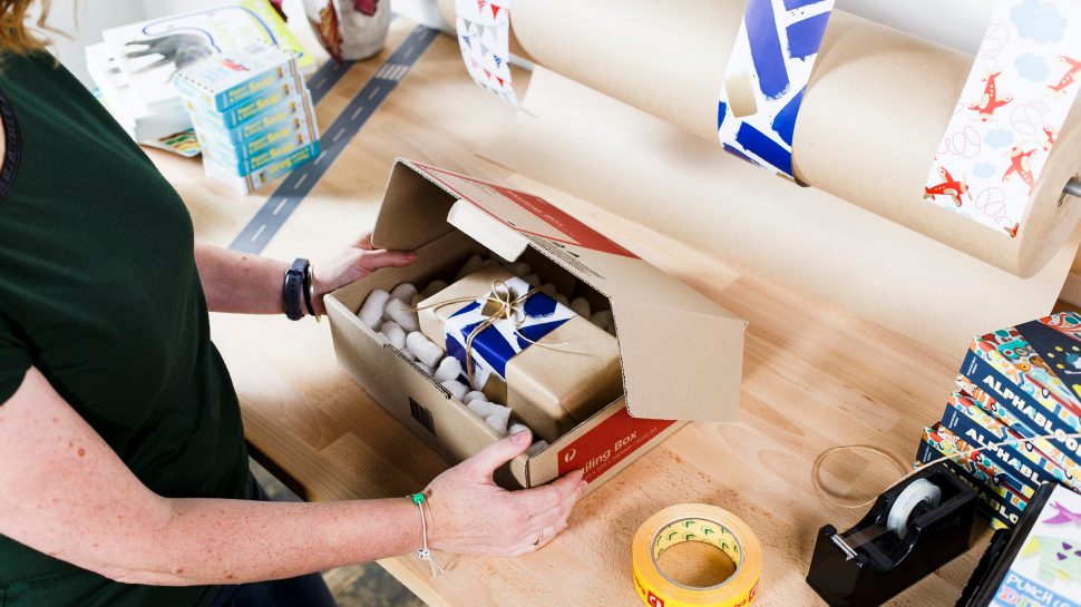 Looking down on a work-bench, Helle is packing an online order in an Australia Post mailing box. To her left and right is stock, ribbon, tape and wrapping paper.