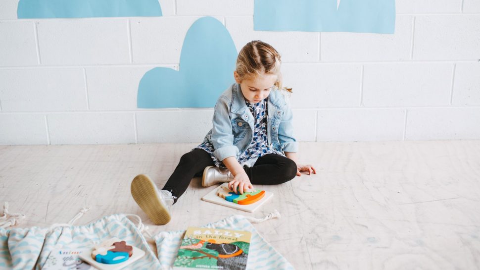 Photograph of a young girl sitting on the floor playing with a toy and with Honey & Co Club canvas bags in front of her