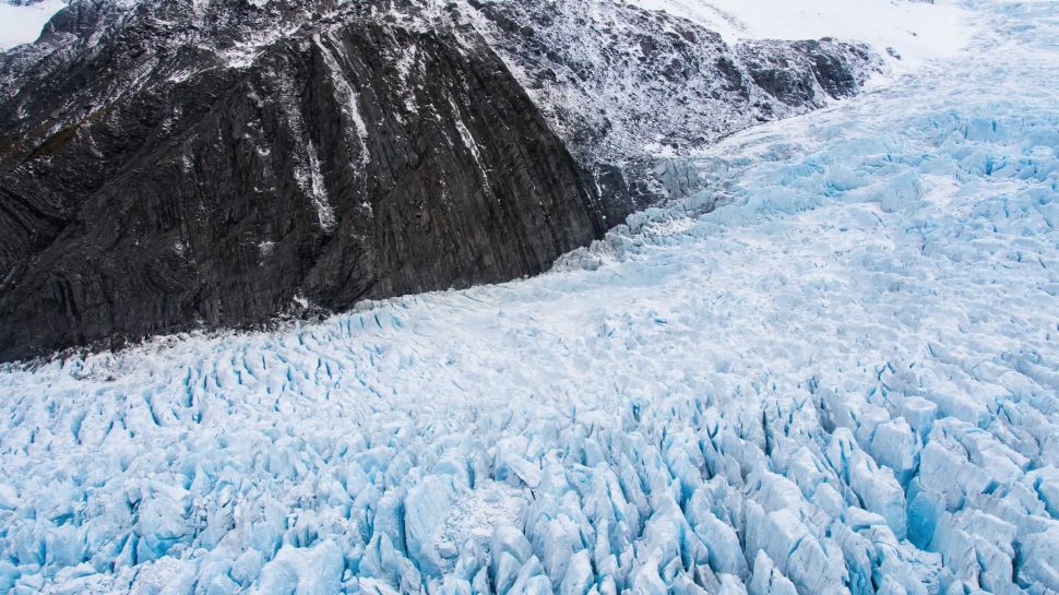 A birds-eye-view of the Fox Glacier in New Zealand. An incredible expanse of light blue, snowy ice forms the glacier, it takes up most of the scene. The ice is rugged with many deep dips on its surface. It travels uphill on the right side of the image. On the left and in the background, the ice joins to a large, dark grey rock on the side of a mountain.